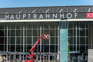 Zwei Personen stehen auf einem Lastenkran und reinigen die Fenster am Eingangsportal des Kölner Hauptbahnhofes. Tauben fliegen durch die Szene Zwei Personen stehen auf einem Lastenkran und reinigen die Fenster am Eingangsportal des Kölner Hauptbahnhofes. Tauben fliegen durch die Szene